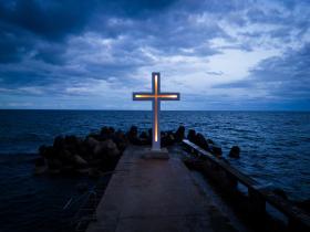 A large Christian cross stands at the edge of a pier against a dramatic sky and sea, seen from above